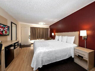 Hotel room with a king-sized bed, red accent wall, mounted tv displaying football, and a small kitchenette.