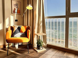 A cozy sea-view room with a bright orange armchair, large windows overlooking the ocean, and modern decor.