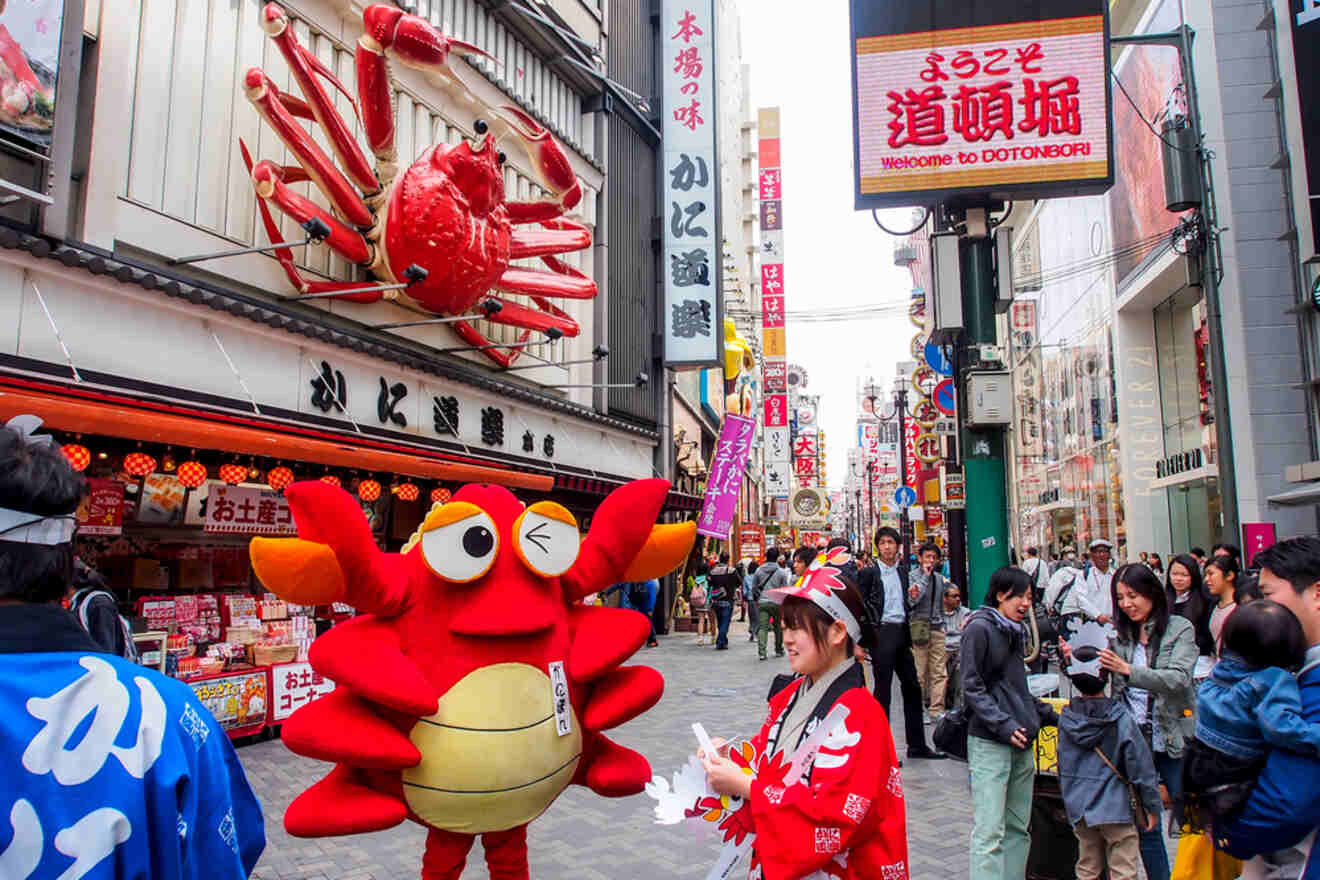 A mascot dressed as a red crab entertaining a crowd on a busy shopping street in osaka, japan, with multiple colorful signs and advertisements.