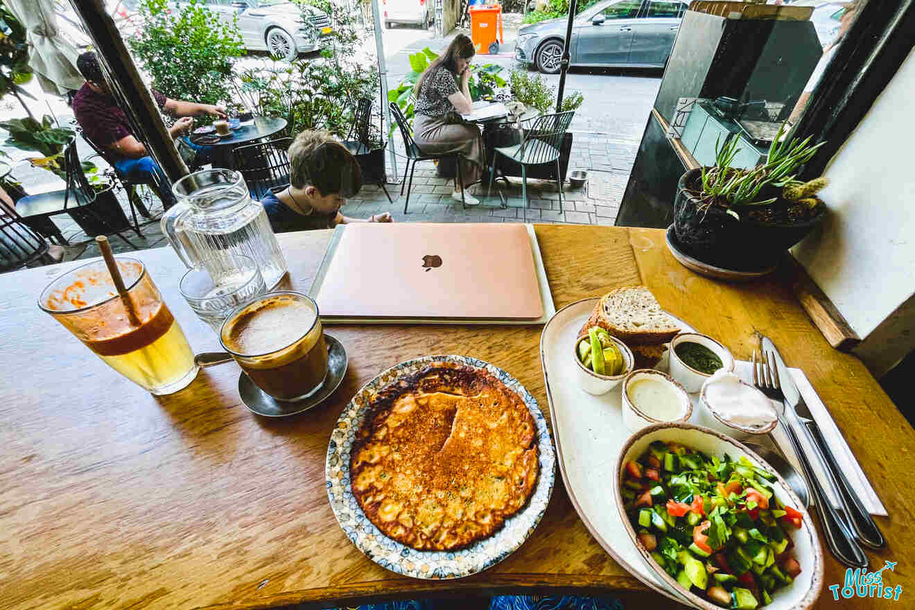 View from a café table with a laptop, a pancake, a coffee, and a salad, overlooking a street-side dining area.