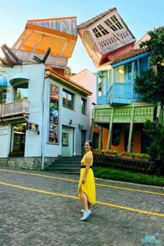 The writer of the post in a yellow dress strolling past Tbilisi's leaning clock tower and colorful buildings, capturing a whimsical and vibrant street scene