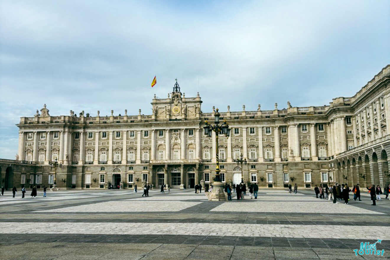 The majestic Royal Palace of Madrid on a cloudy day, with tourists dotting the expansive plaza in front of the palace's classical architecture.