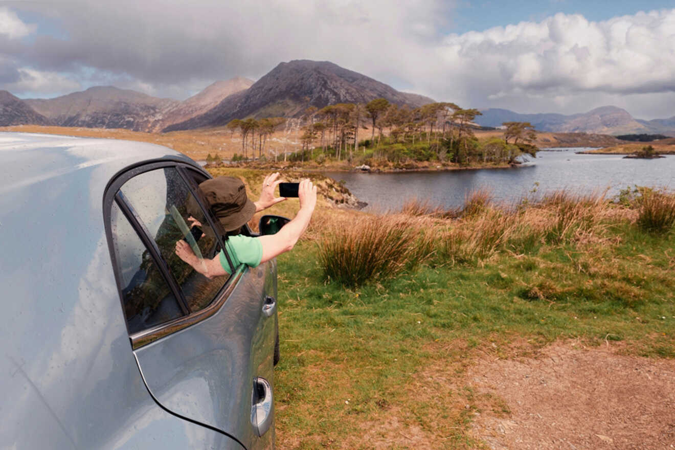 A person leaning out of a car window taking a photo with their smartphone, framing a picturesque view of a lake and mountains, highlighting the beauty of Irish landscapes