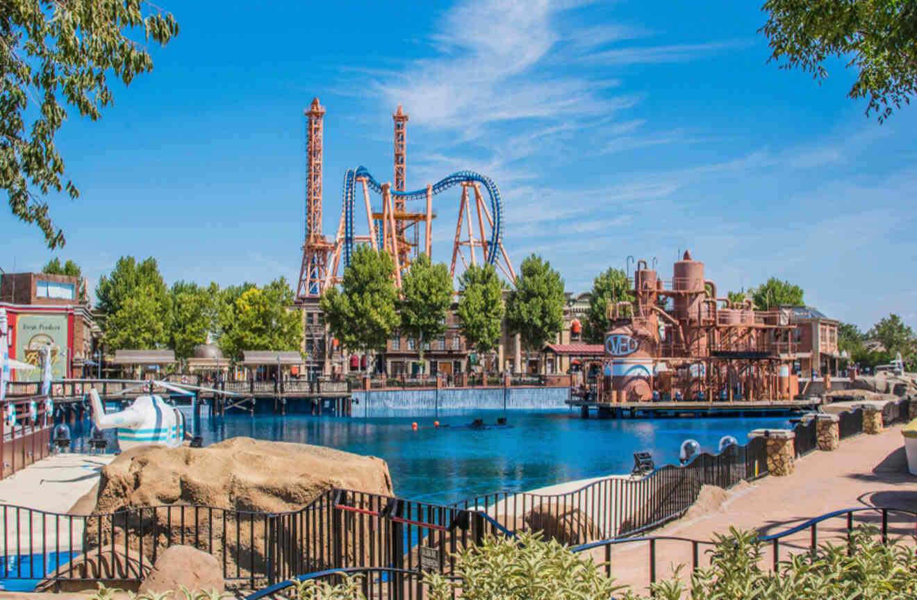 A view of Parque Warner Madrid, an amusement park with a large lake in the foreground and roller coasters against a clear blue sky.