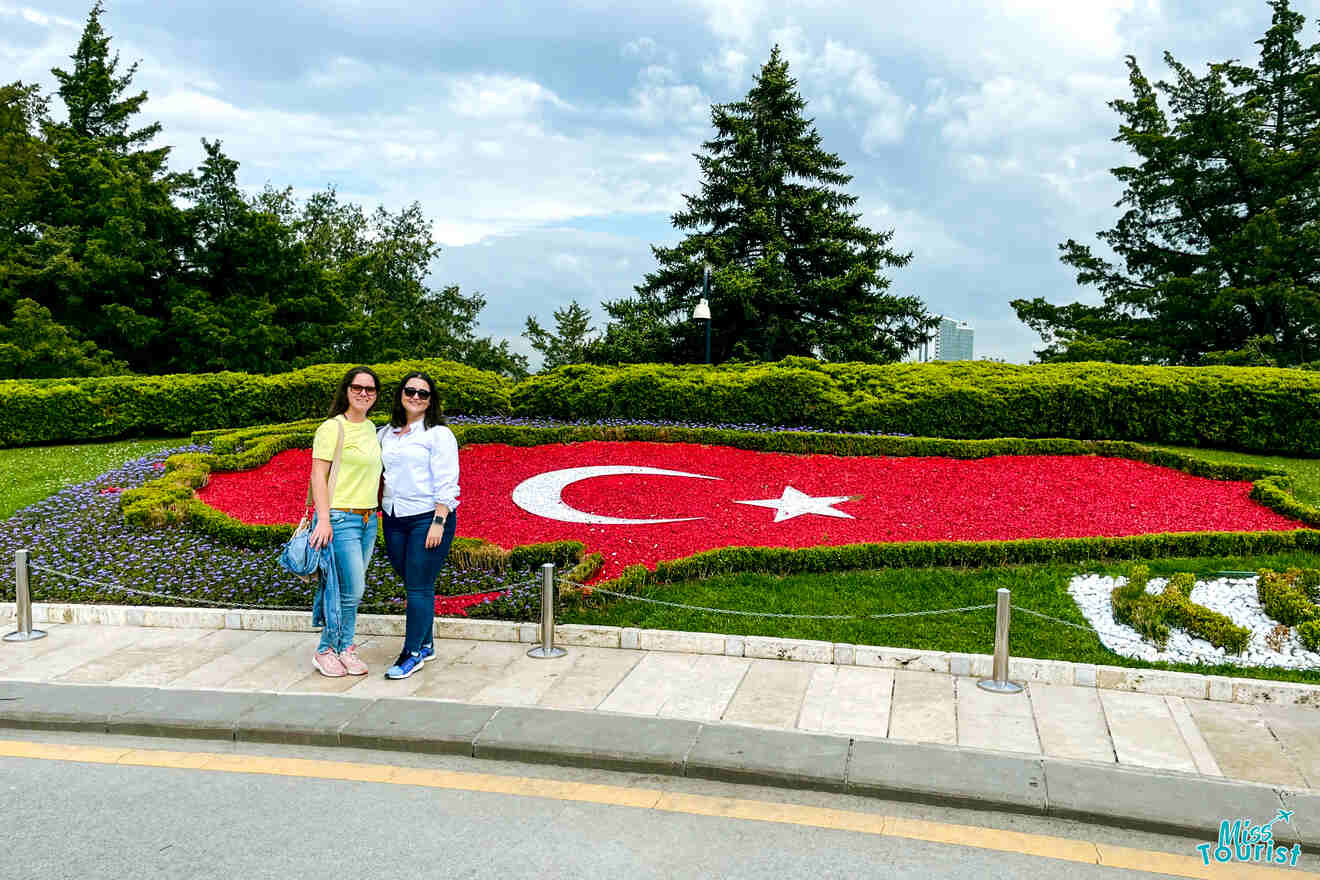 the author of the post with a friend posing in front of a large floral display shaped like the turkish flag in a park.