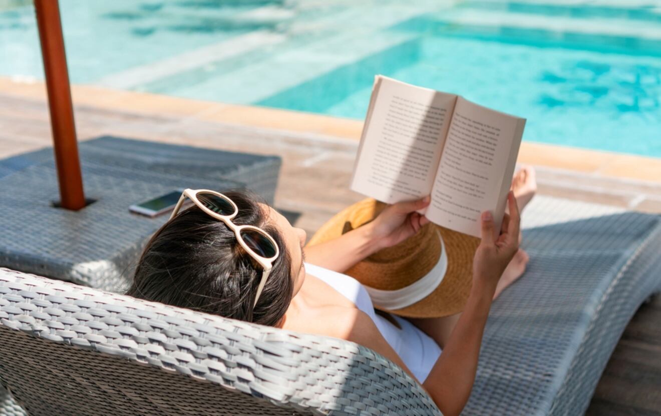 Person lounging on a wicker sunbed reading a book by a tranquil swimming pool, with a smartphone and a straw hat nearby