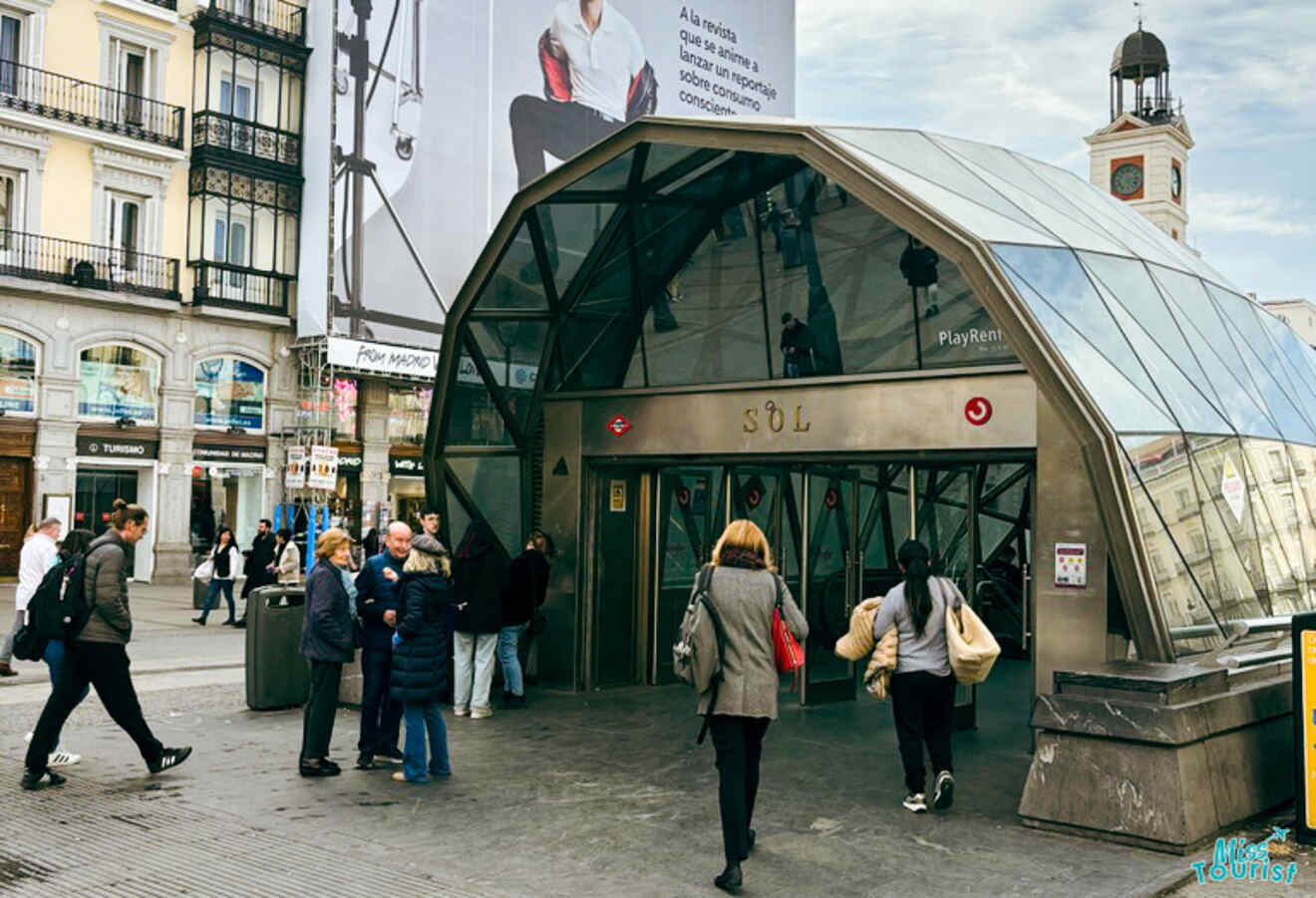 Pedestrians entering and exiting the modern glass-and-metal Sol metro station in Madrid, with the bustling city life around