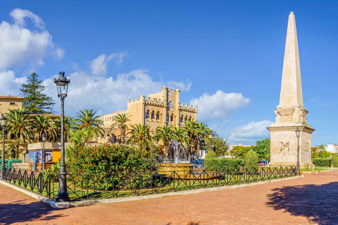 The historic plaza in Ciutadella de Menorca, with a tall obelisk monument, lush gardens, and a medieval sandstone building under a bright blue sk