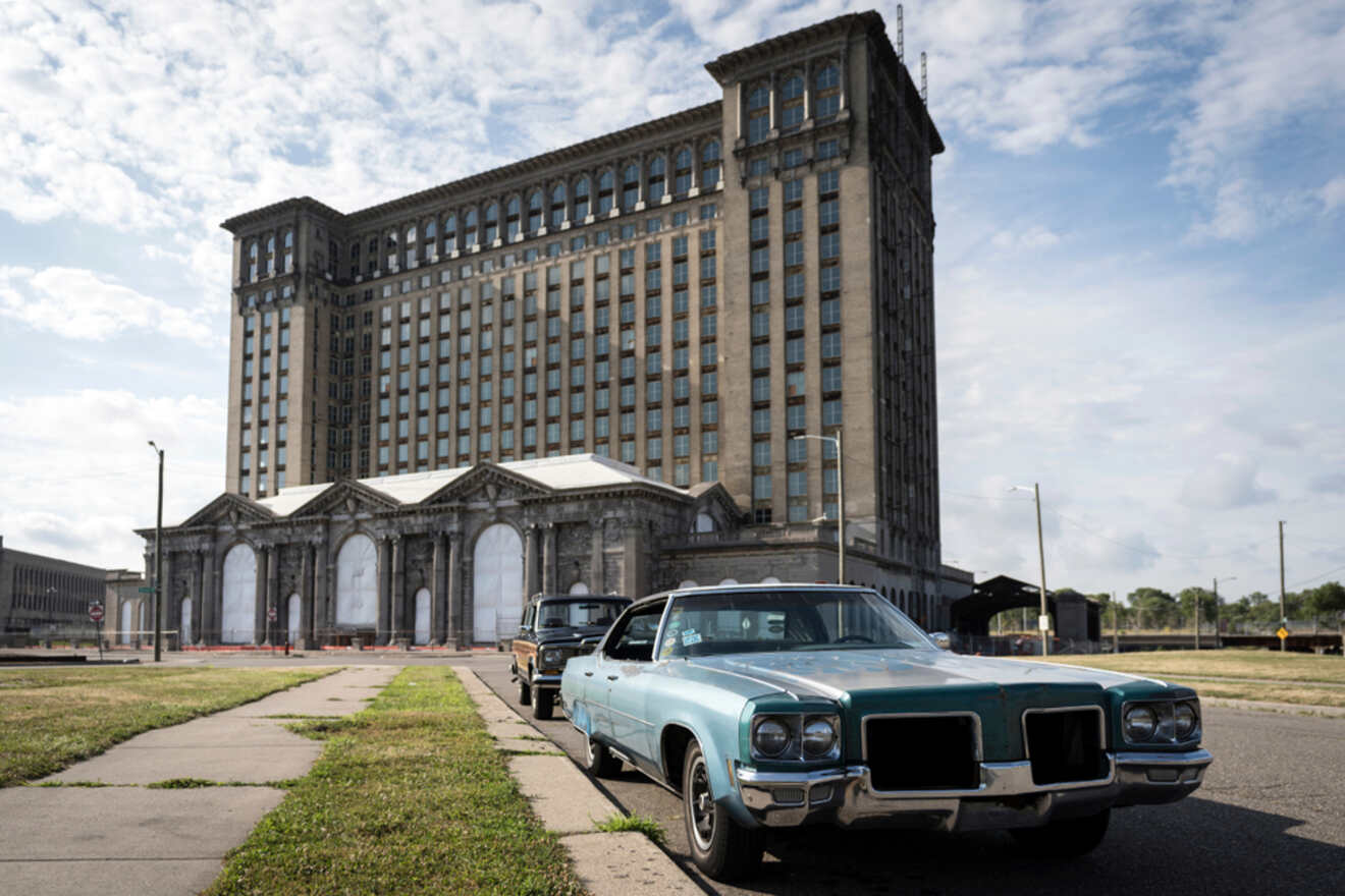 The historic and abandoned Michigan Central Station in Detroit framed by a clear blue sky, with a vintage teal car parked out front.