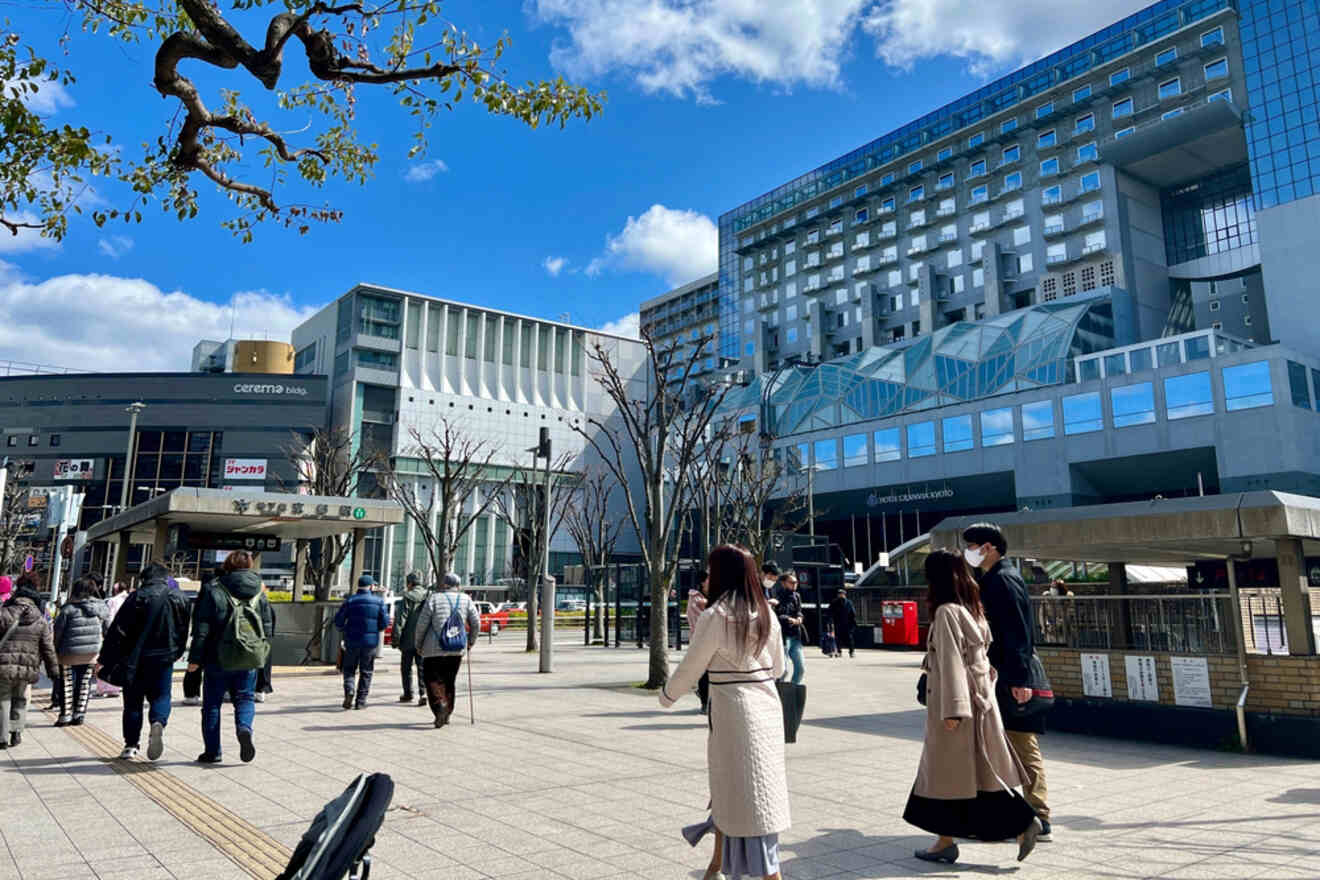 A bustling area near Kyoto Station with modern architecture, people walking, and bare trees against a backdrop of blue sky.