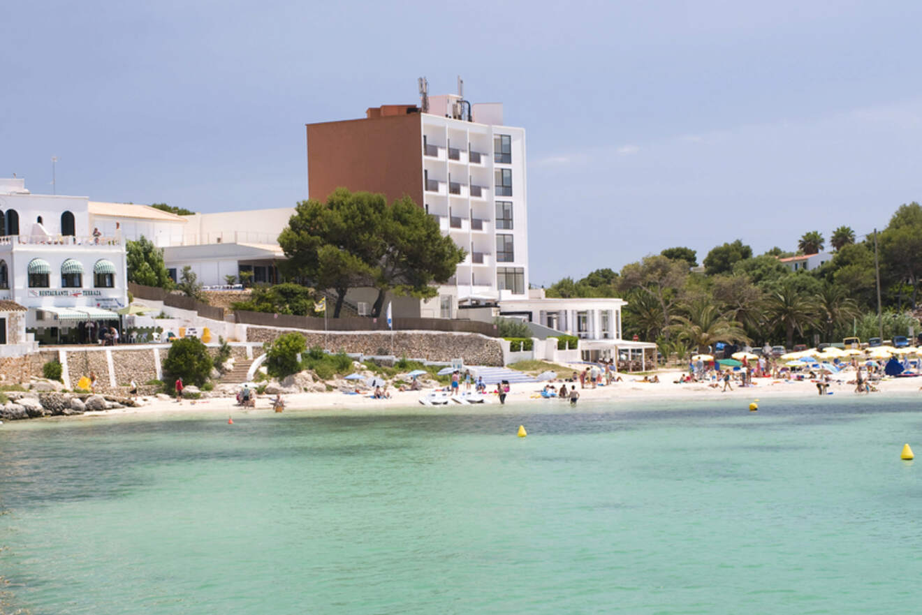 The tranquil beach of Cala Santandria in Menorca, with crystal clear shallow waters, sunbathers on the white sand, and a backdrop of modern white buildings and greenery