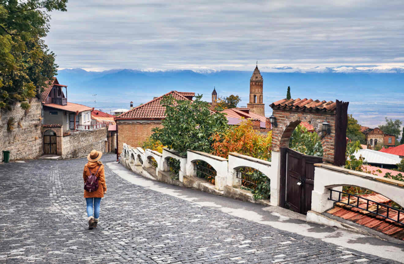 A solitary figure walks down a cobblestone street in Sighnaghi, past historic buildings with a backdrop of distant snow-capped mountains, evoking a sense of wanderlust