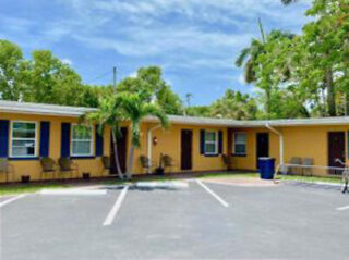 A colorful motel with yellow walls and blue trim, featuring palm trees and parking spaces in front.