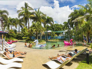 People relaxing on sun loungers and enjoying the pool on a sunny day, surrounded by lush greenery and palm trees.