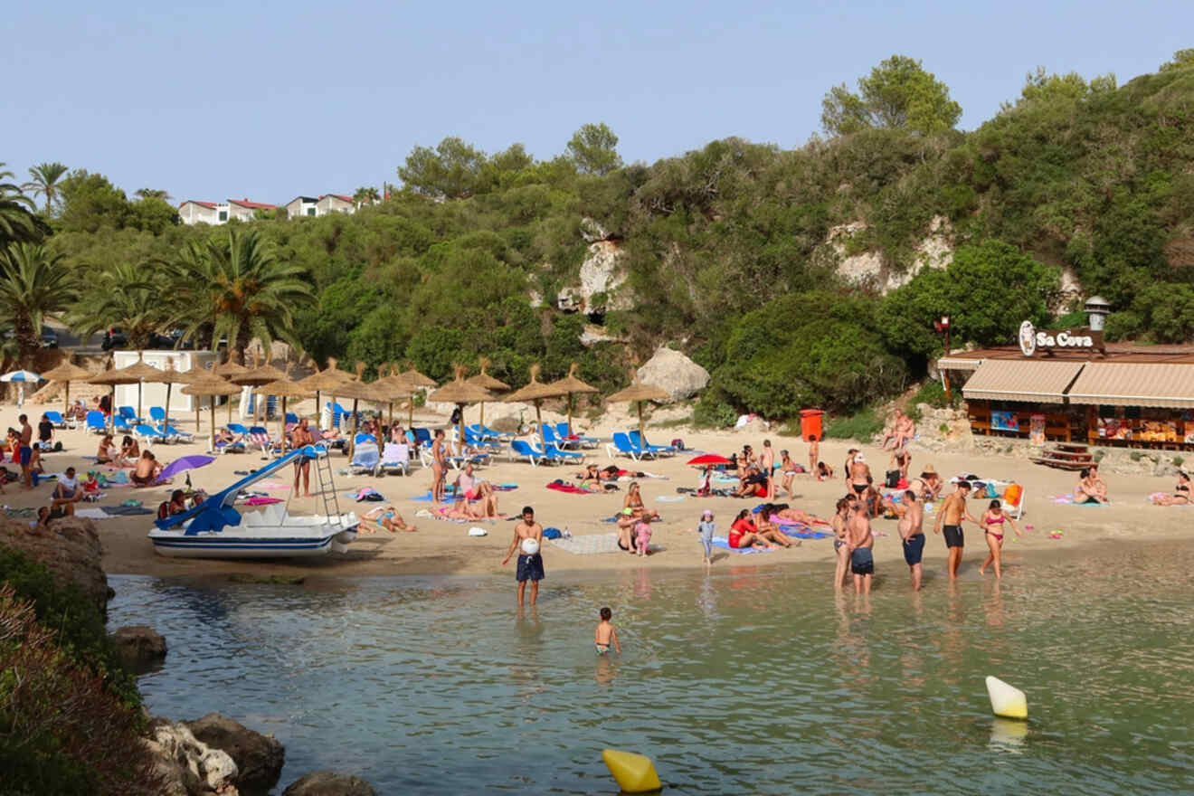 Vibrant beach scene at Los Delfines & Cala en Blanes with holiday-goers enjoying the sandy shore, straw umbrellas, and a beach bar nestled against a lush green hillside