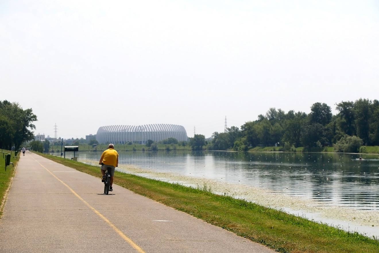 A person in a yellow shirt riding a bicycle on a paved path next to a calm river, with a unique, ribbed structure in the distance, on a hazy day.