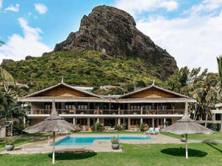 A luxury resort with a thatched-roof structure and a swimming pool, situated at the base of a tall, rugged mountain.