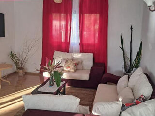 Cozy living room with a red sofa, two white armchairs, a glass coffee table, red curtains, and several green plants.