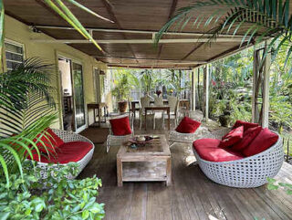 Covered outdoor patio with red cushioned chairs, a wooden coffee table, and lush greenery surrounding the area.