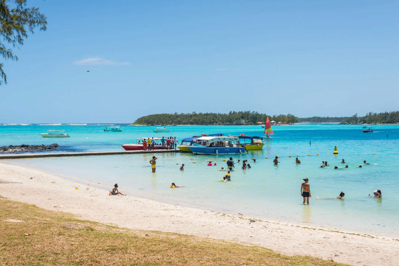 Sandy beach in Mauritius with locals and tourists enjoying the clear shallow waters near moored boats and a view of a small island
