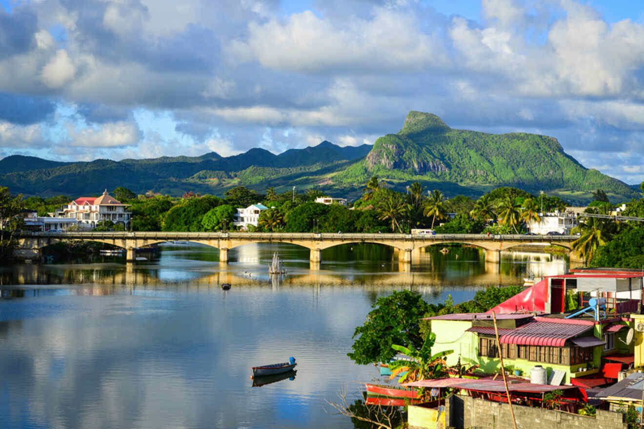 Scenic view of Mahebourg with the tranquil Rivière La Chaux, traditional boats, and a bridge against a mountainous horizon in Mauritius