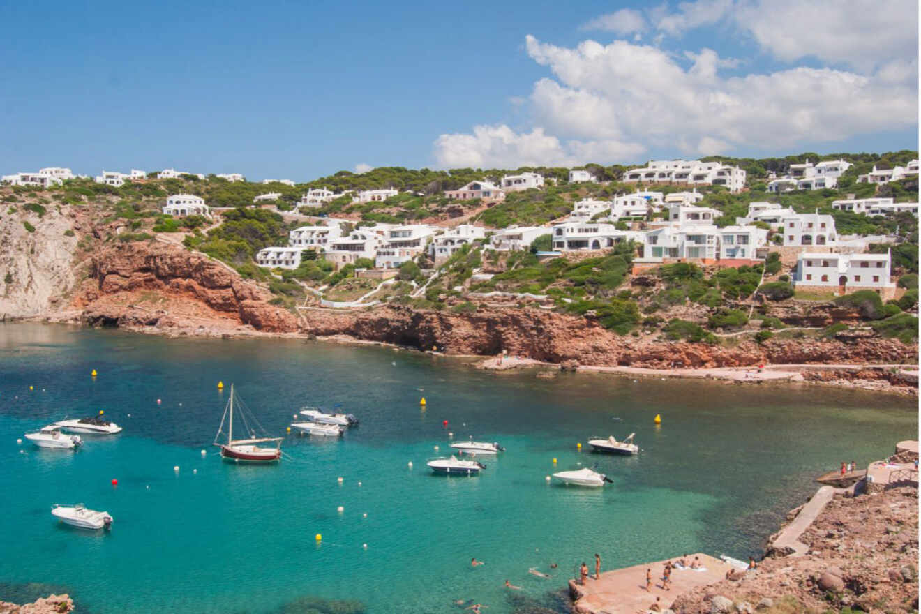 Breathtaking view of Cala Morell with white villas perched atop red cliffs overlooking a tranquil cove with crystal-clear waters and moored boats in Menorca