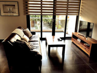Comfortable living area with a plush dark sofa, wood finish entertainment unit, and natural light filtering through blinds