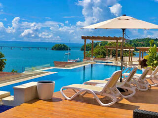 A luxurious infinity pool with a sweeping ocean view, white lounge chairs under an umbrella, and a clear blue sky.