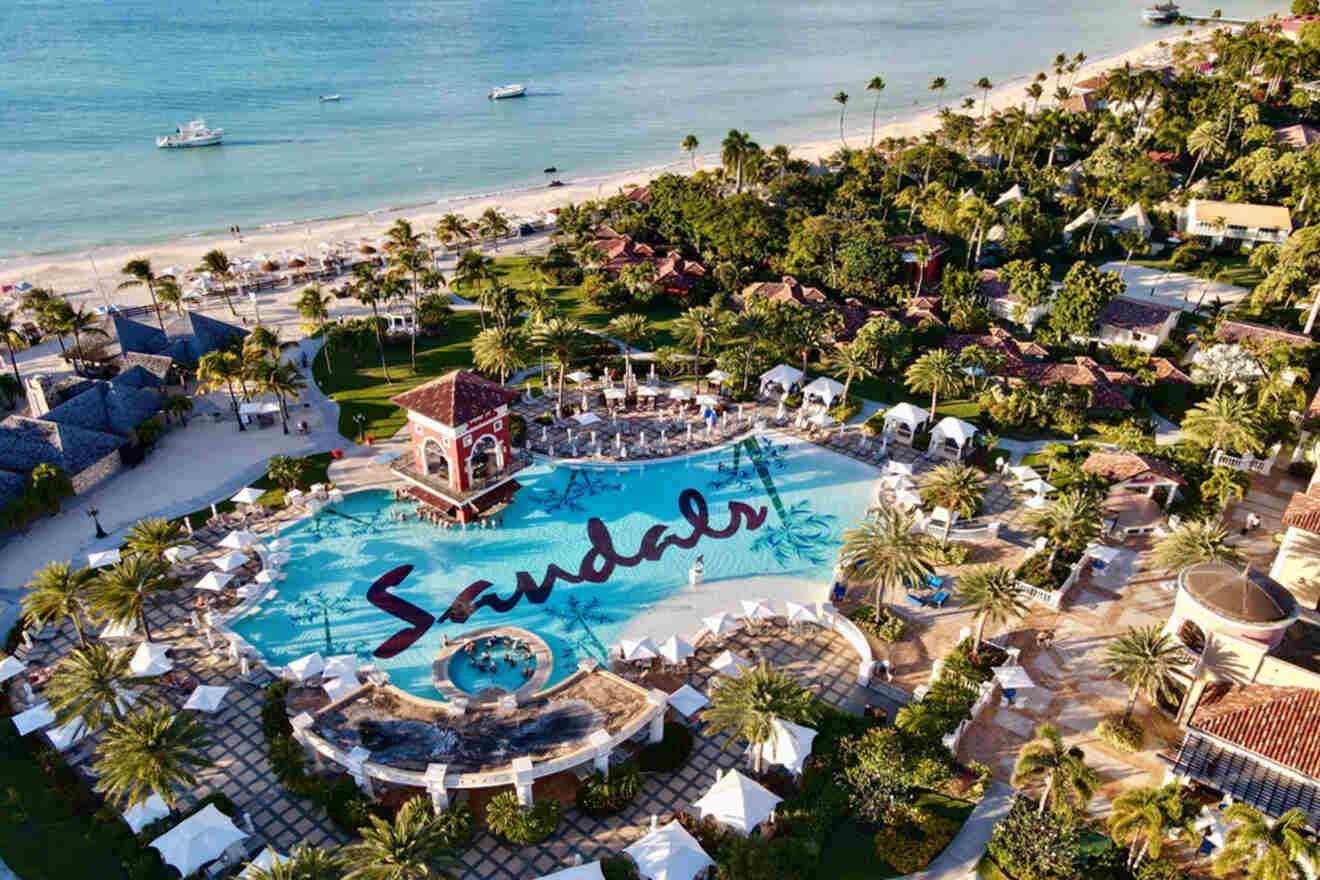Aerial view of a sandals resort with a large pool, surrounding palm trees, beachfront, and boats in the sea.