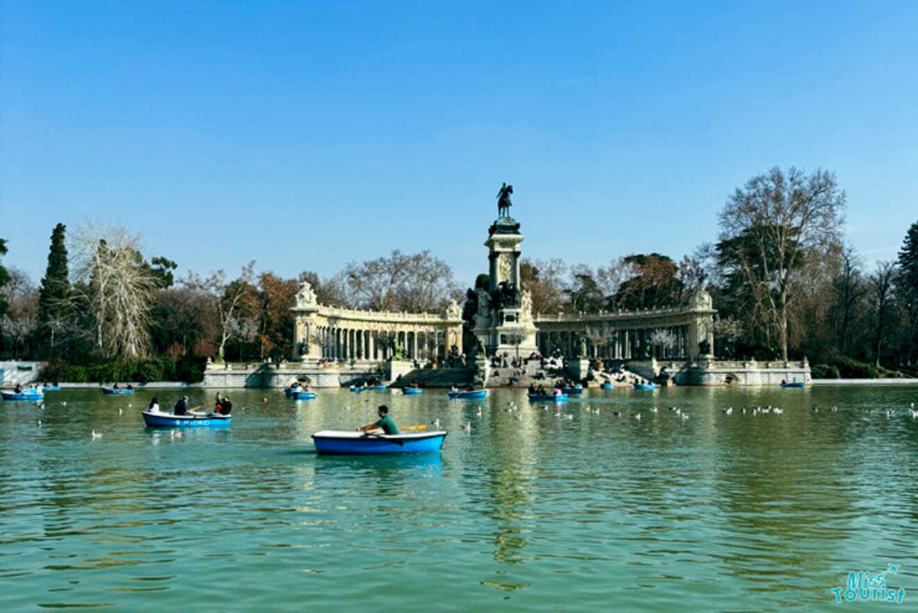 Visitors leisurely rowing boats on the peaceful green waters of the Retiro Park lake, with the Monument to Alfonso XII in the background on a sunny day.
