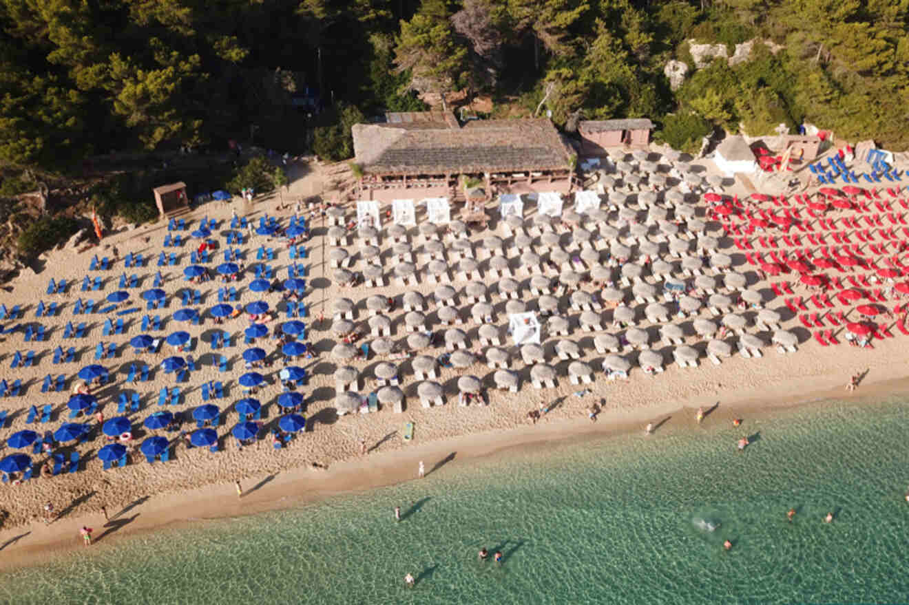 Aerial view of a crowded beach with rows of blue and red umbrellas, adjacent to clear turquoise waters and surrounded by trees.