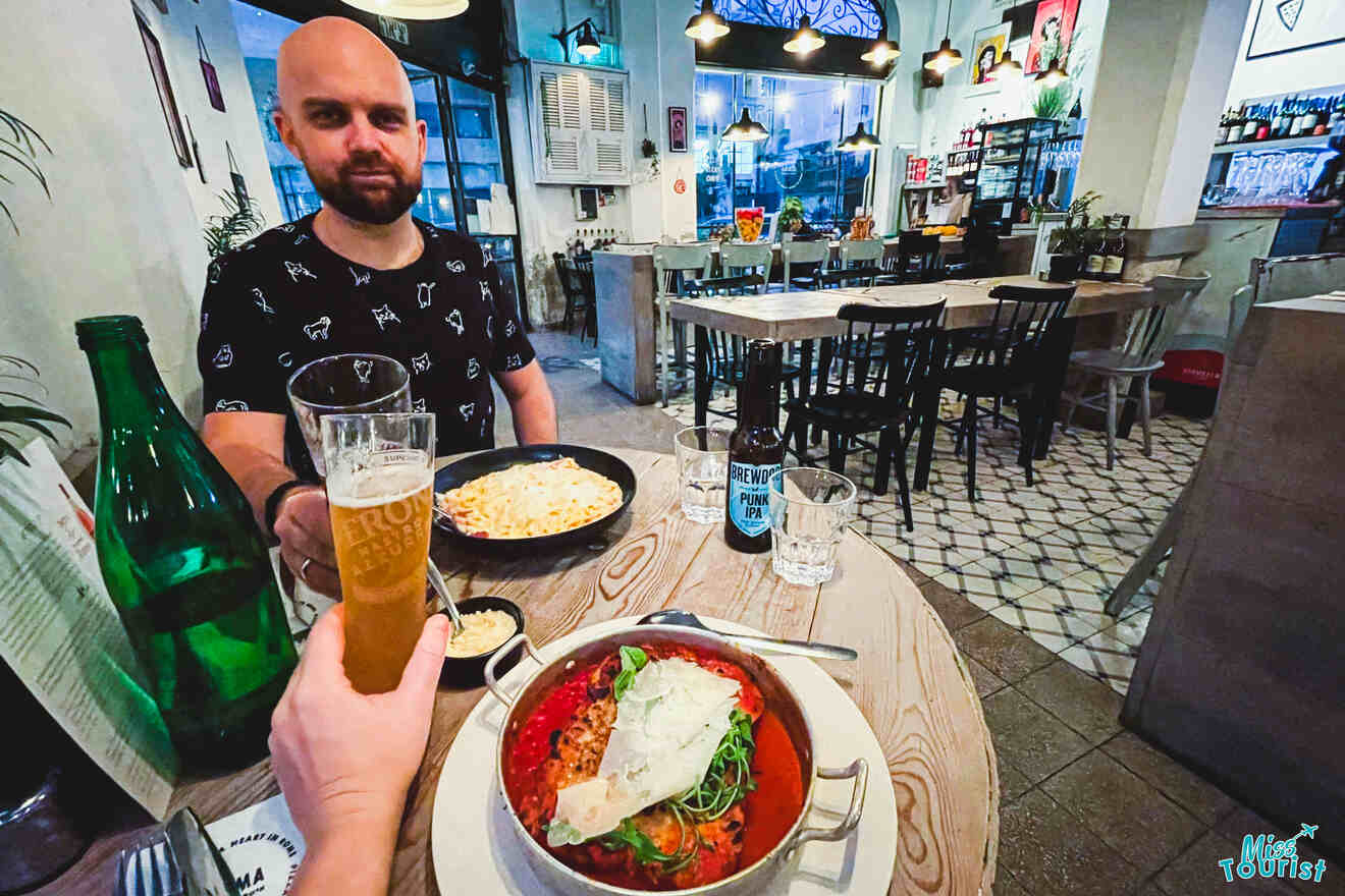 A man at a restaurant table holding a beer, with plates of food including soup and pasta, in a casually decorated dining space.