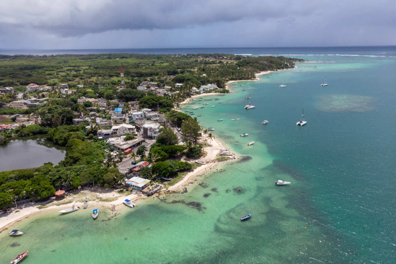 Aerial view of a coastal village in Mauritius, with a mix of dense vegetation and scattered buildings, flanking a tranquil turquoise sea