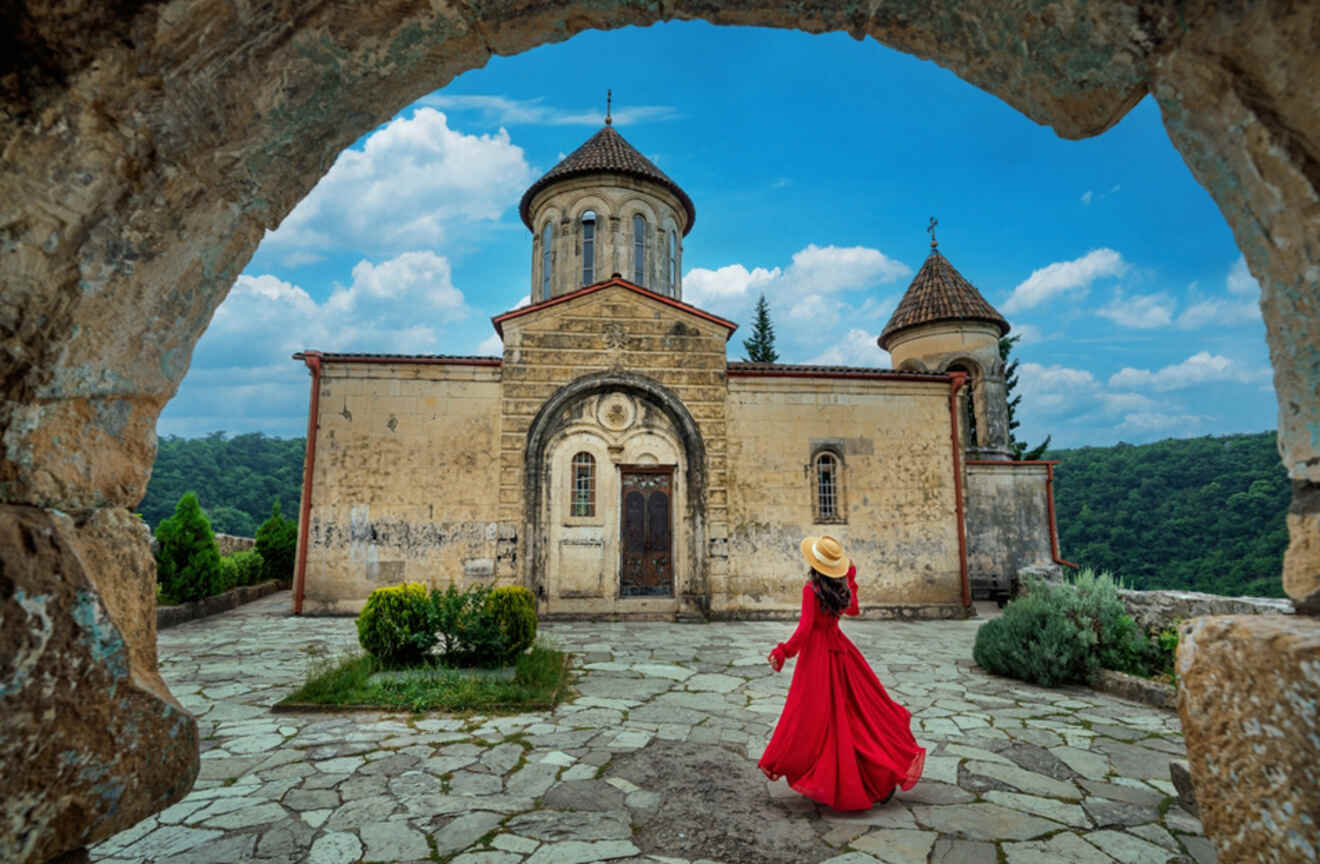 A woman in a striking red dress and straw hat stands by an ancient stone archway, framing the historic Motsameta Monastery against a backdrop of lush greenery and a clear sky.