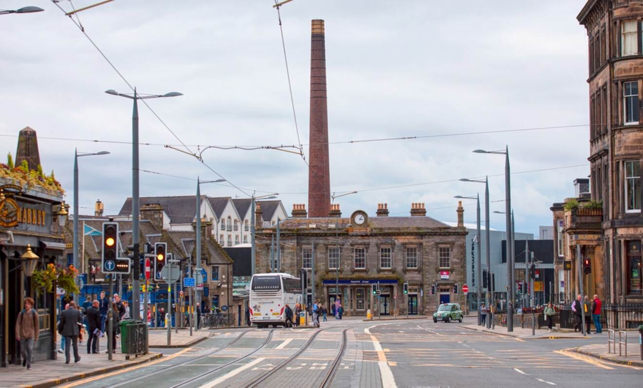 Urban street scene in Edinburgh with historical buildings, a prominent brick chimney, and tram tracks, bustling with pedestrians and vehicles