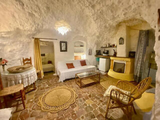 Interior of a cozy, cave-style room with white stone walls, a small kitchen area, rustic furniture, and a round woven rug on the tiled floor.