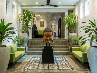 A spacious lobby with a large buddha head sculpture, flanked by green sofas and potted plants, all under a modern chandelier.