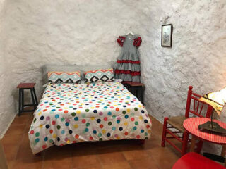 A cozy, rustic bedroom with a polka dot bedspread, a flamenco dress hanging on the wall, and simple wooden furniture.