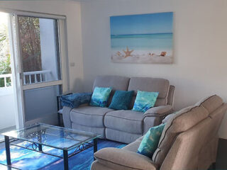 A modern living room with a gray sofa, turquoise cushions, a glass coffee table, and a beach-themed painting above the sofa. a window with a garden view is on the left.