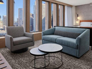 Hotel room with a cozy sitting area, showcasing a gray sofa, armchair, and circular coffee tables, with city views