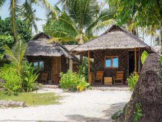 Two traditional thatched-roof huts surrounded by lush palm trees on a sandy path, typical of a tropical island setting.