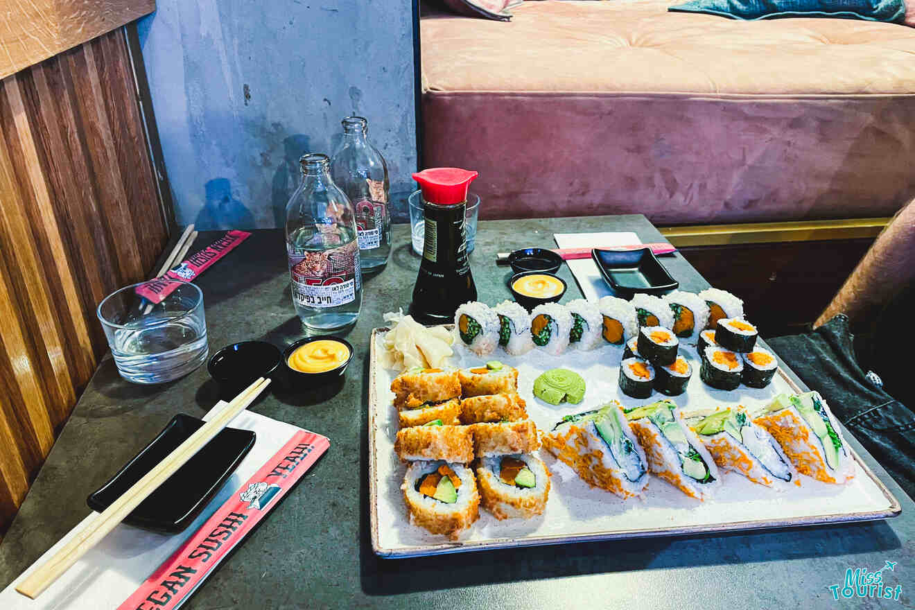 A tray of assorted sushi rolls served with soy sauce, wasabi, and ginger on a table in a restaurant, with chopsticks and beverages alongside.