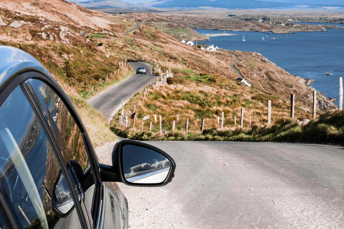 Curved road along a coastal hillside with a car's side mirror reflecting the scene, capturing the essence of a scenic drive on Ireland's rugged coastline