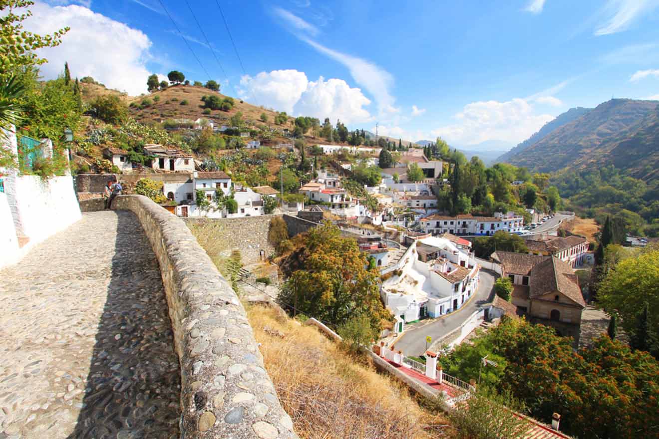 View of a traditional white village nestled in the hills, photographed from atop an old stone bridge on a sunny day.