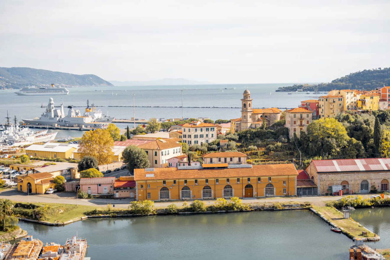 Aerial view of La Spezia's coastline, featuring historic buildings, docks, and a clear view out to the sea with vessels and rolling hills in the background.