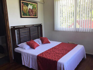 A neatly made bed with a red and white bedspread in a room with wooden floors, a floral painting above the bed, and sunlight filtering through vertical blinds.