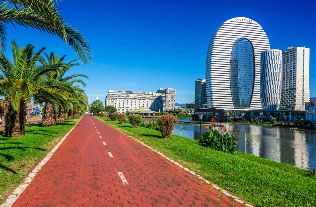 A vibrant walkway lined with palm trees leading towards modern, curved buildings by a tranquil waterway in Batumi, reflecting a blend of natural beauty and contemporary urban design