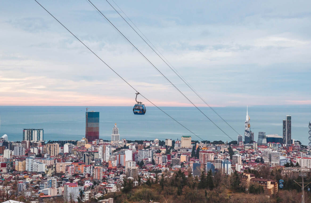 A cable car glides over Batumi, offering panoramic views of the cityscape with diverse architecture, nestled between the sea and the rolling hills at dusk