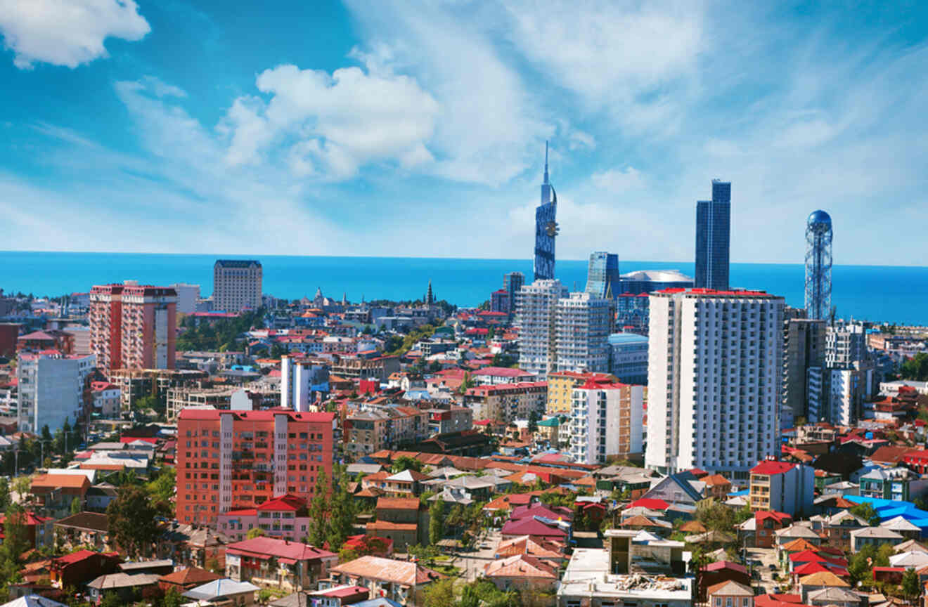 A bustling cityscape of Batumi, featuring a mix of historic and modern buildings, with the Black Sea in the backdrop under a sky dotted with fluffy clouds