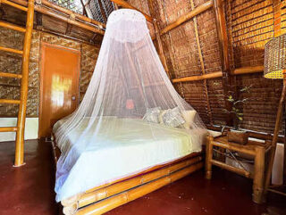 A cozy bedroom featuring a bed draped with a white mosquito net, bamboo walls and furniture, and warm lighting.